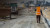 工人身穿橙色安全背心，手持 Trimble GNSS 接收机，正在重铺路面。