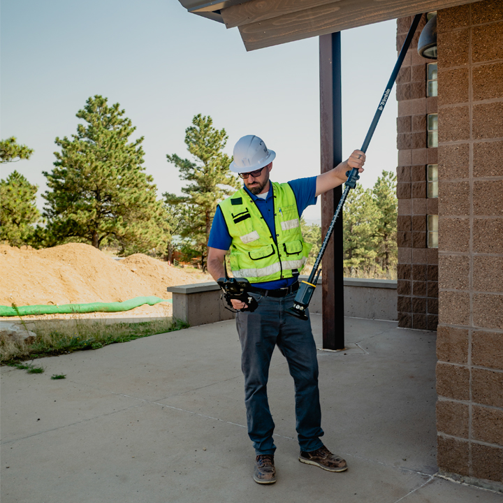 A surveyor in a hard hat and safety vest holds the ST30 smart target upside down against the underside of a building overhang whilst looking at his survey controller.