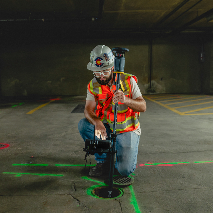 A surveyor in a high-visibility vest uses a Trimble ST30 smart target in a parking garage. He is kneeling while operating survey equipment.