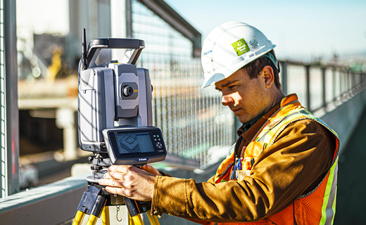 Surveyor on construction site next to a fence looking at a Trimble total station.