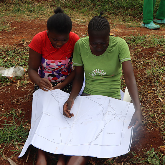 Two women sitting on the ground on a land parcel in Africa, reviewing a surveying document together.