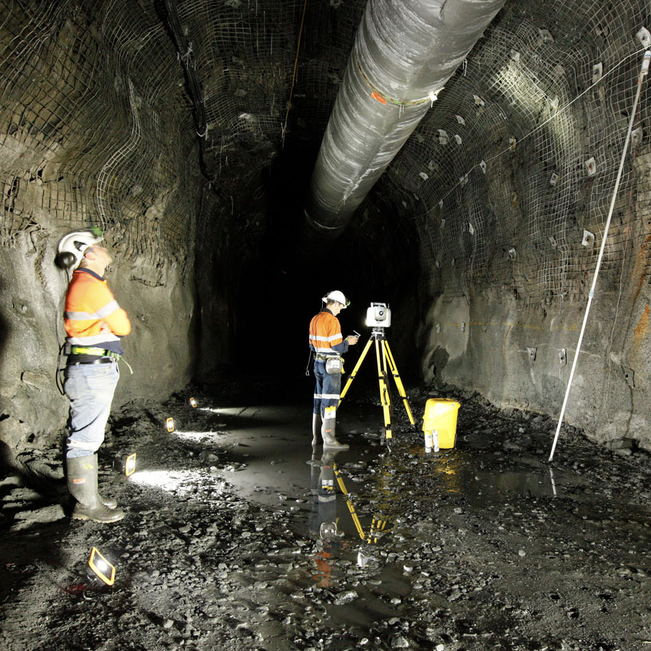 Two workers in high-visibility vests using a Trimble SX12 total station and scanner set up on a tripod