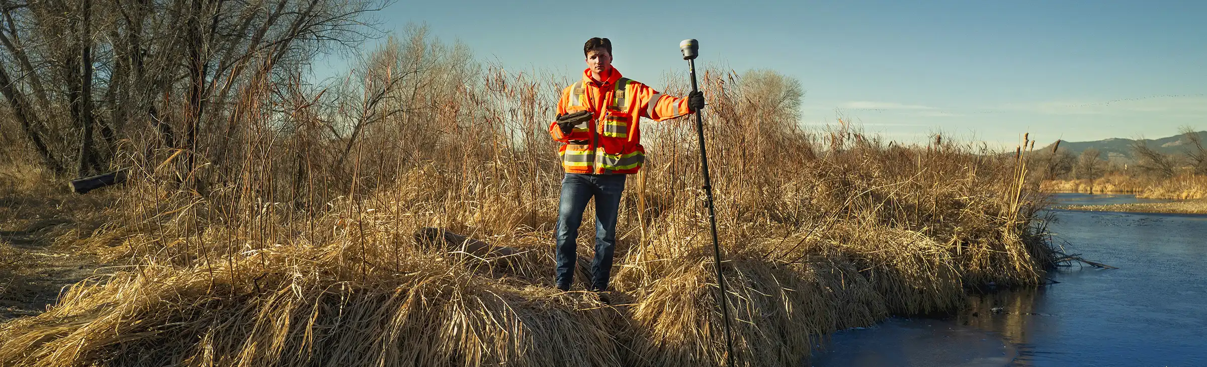 一名测量员站在河边，一手持 Trimble T70 平板电脑，另一手持 R12i 接收机。