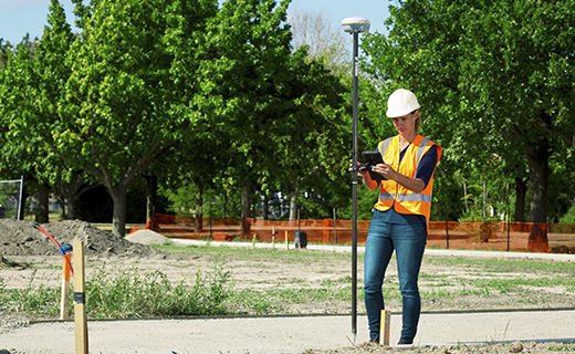 Topógrafo con casco de pie en una obra de construcción de una subdivisión residencial, que sostiene un sistema móvil Trimble GNSS y realiza mediciones.
