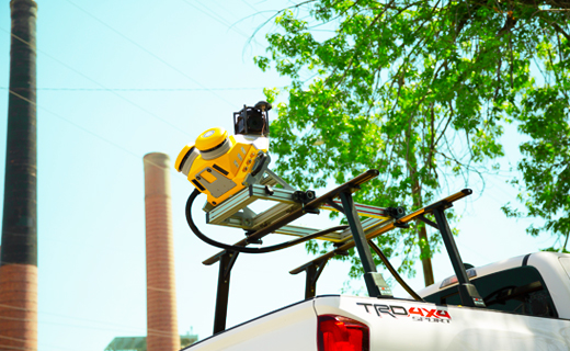 Trimble MX50 mobile mapping system mounted on top of a white SUV vehicle next to green tree branches and tall red brick chimney structures.