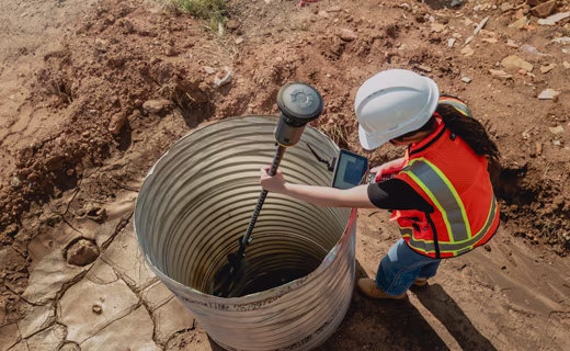 A contractor in a hard hat and safety vest stands over a large metal culvert pipe embedded in the dirt. She is holding the ST30 smart target inside the culvert and looking at her survey controller.