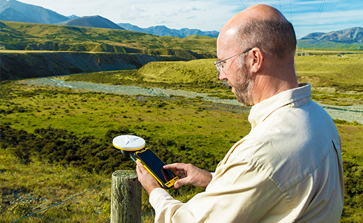 Homme debout près d’un poteau de clôture dans un paysage rural, tenant un accessoire Trimble Catalyst Handle auquel est fixé un dispositif portable Trimble TDC600. 