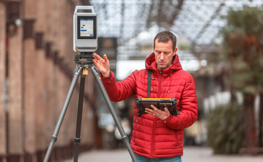 Topógrafo con chaqueta roja de pie junto a un escáner láser Trimble X12 mientras mira la tableta Trimble que tiene en la mano.