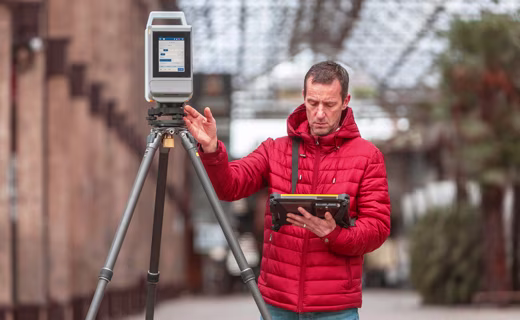 Vermessungsingenieur in roter Jacke steht neben einem Trimble X12 Laserscanner und blickt auf das Trimble Tablet in seiner Hand.