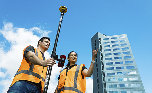 Man holding a survey pole with Trimble DA2 receiver on top. He and a colleague are wearig high visibility vests and standing next to a tall city building.