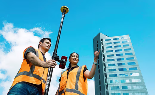 Homme tenant une canne de levé surmontée d'un récepteur Trimble DA2. Lui et sa collègue portent des gilets à haute visibilité et se tiennent à côté d'un grand building.