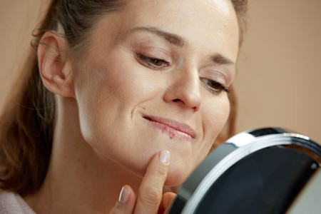 Image of close-up of a woman looking in a mirror and touching her chin, appearing to examine a cold sore.