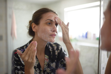 Woman with adult acne applying cleanser on her face in front of a mirror.
