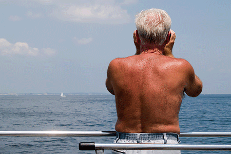 Suntanned back of elderly man that is peering out over blue water. Expresses concepts of sun exposure.