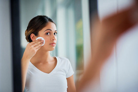 Young woman with acne cleaning her face in front of a mirror
