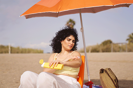 Woman applying sunscreen under an umbrella at the beach.