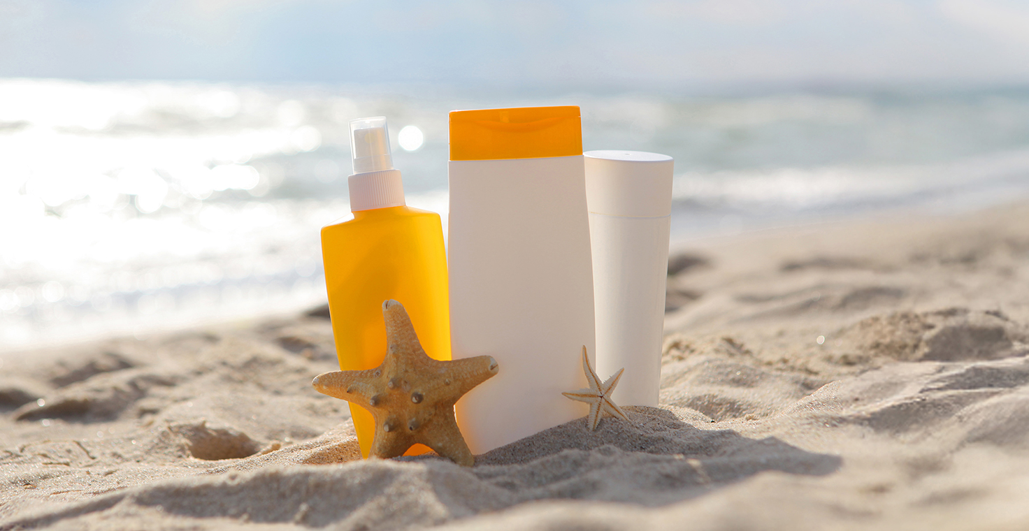 Bottles of sunscreen with starfish on a sandy beach next to glistening ocean water. 
