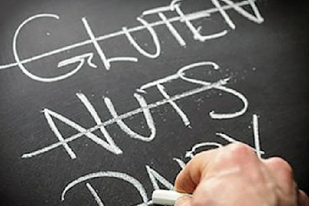 A hand lingers over a blackboard where the words of common food allergens, “gluten” and “nuts,” are crossed out. 