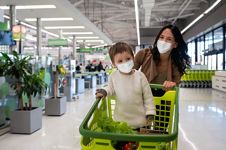 Mother grocery shopping with her toddler at the supermarket wearing face masks and pushing the cart.
