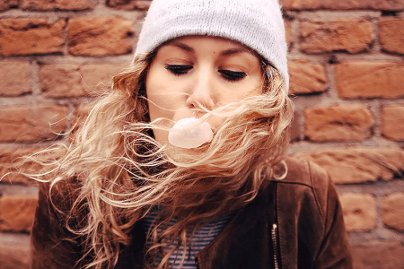 Teen girl blowing a pink bubble gum bubble with a brick wall behind her.