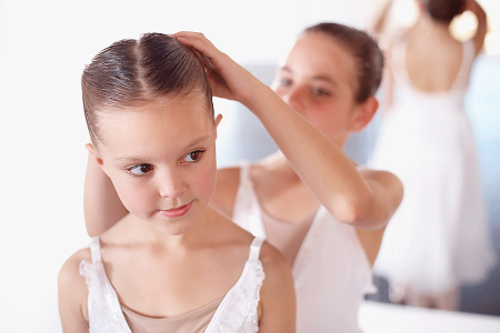 Young ballet dancer pulls another dancer’s hair back in a tight bun.