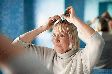 Senior woman applying serum to scalp to prevent hair loss, looking at mirror in bathroom