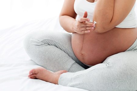 Pregnant person with eczema sits on a bed and rubs lotion on dry skin on their elbow.