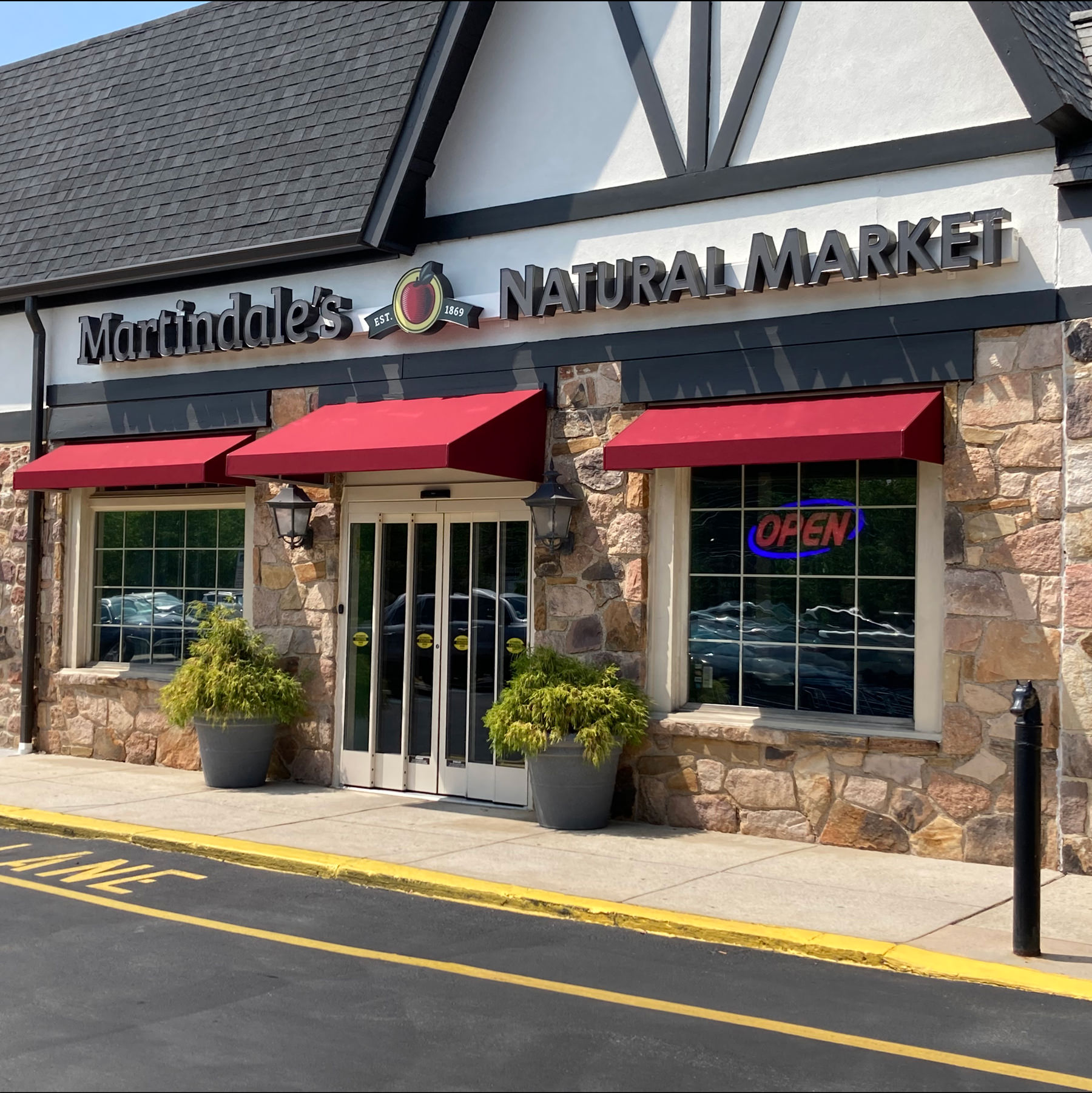 Martindale’s Natural Market storefront with large sign, stone facade, red awnings, two windows, glass doors, and potted yellow plants by the entrance.
