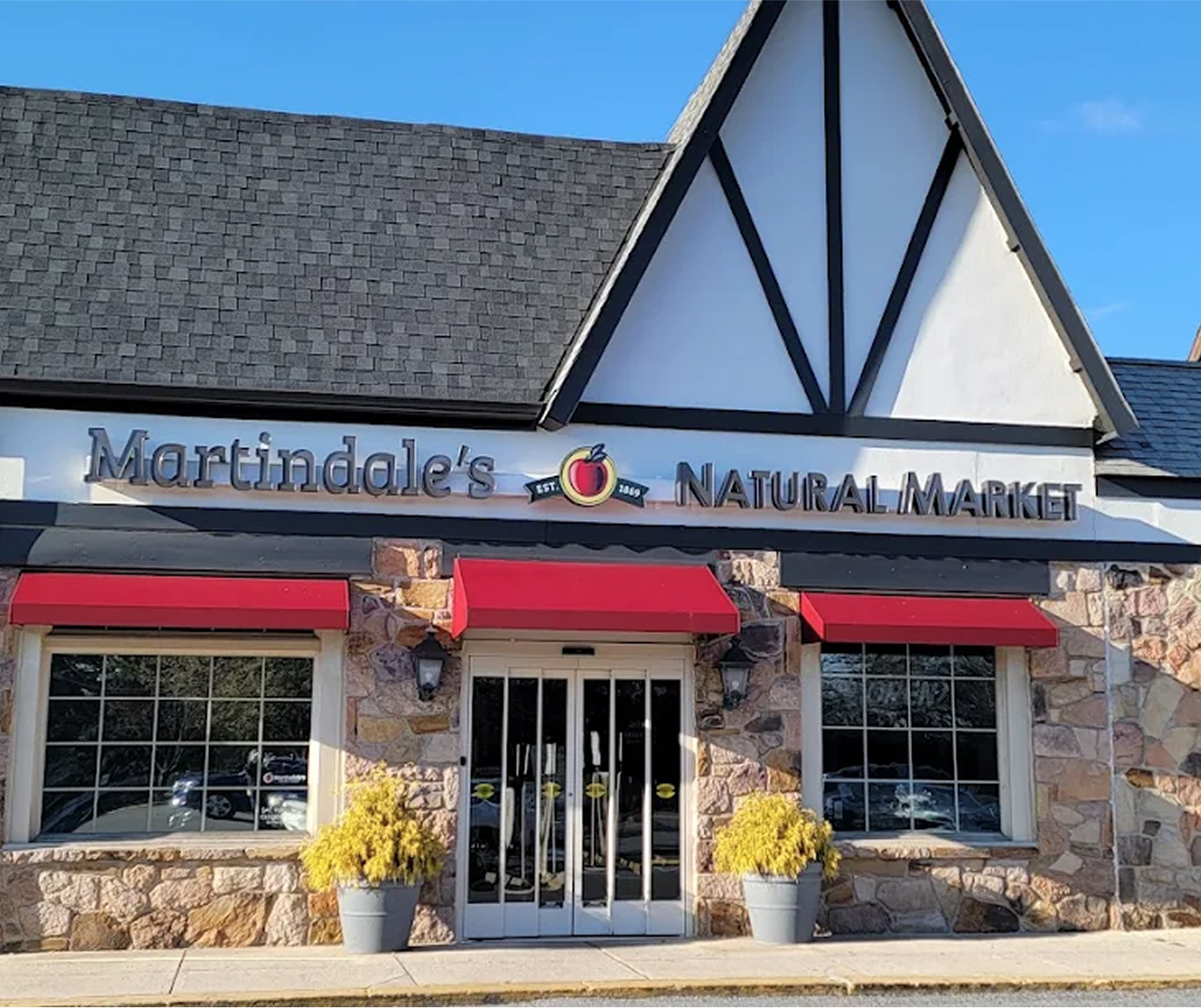Martindale’s Natural Market storefront with large sign, stone facade, red awnings, two windows, glass doors, and potted yellow plants by the entrance.