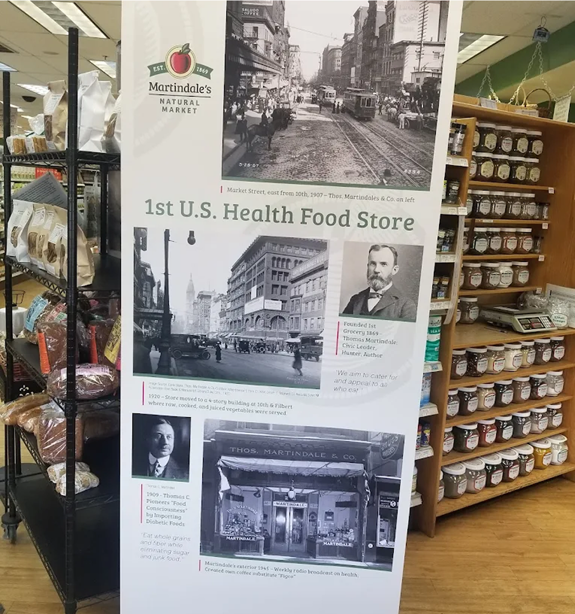 A display board in a grocery store highlights Martindales Natural Market as the 1st U.S. health food store, featuring historical photos and a brief history.