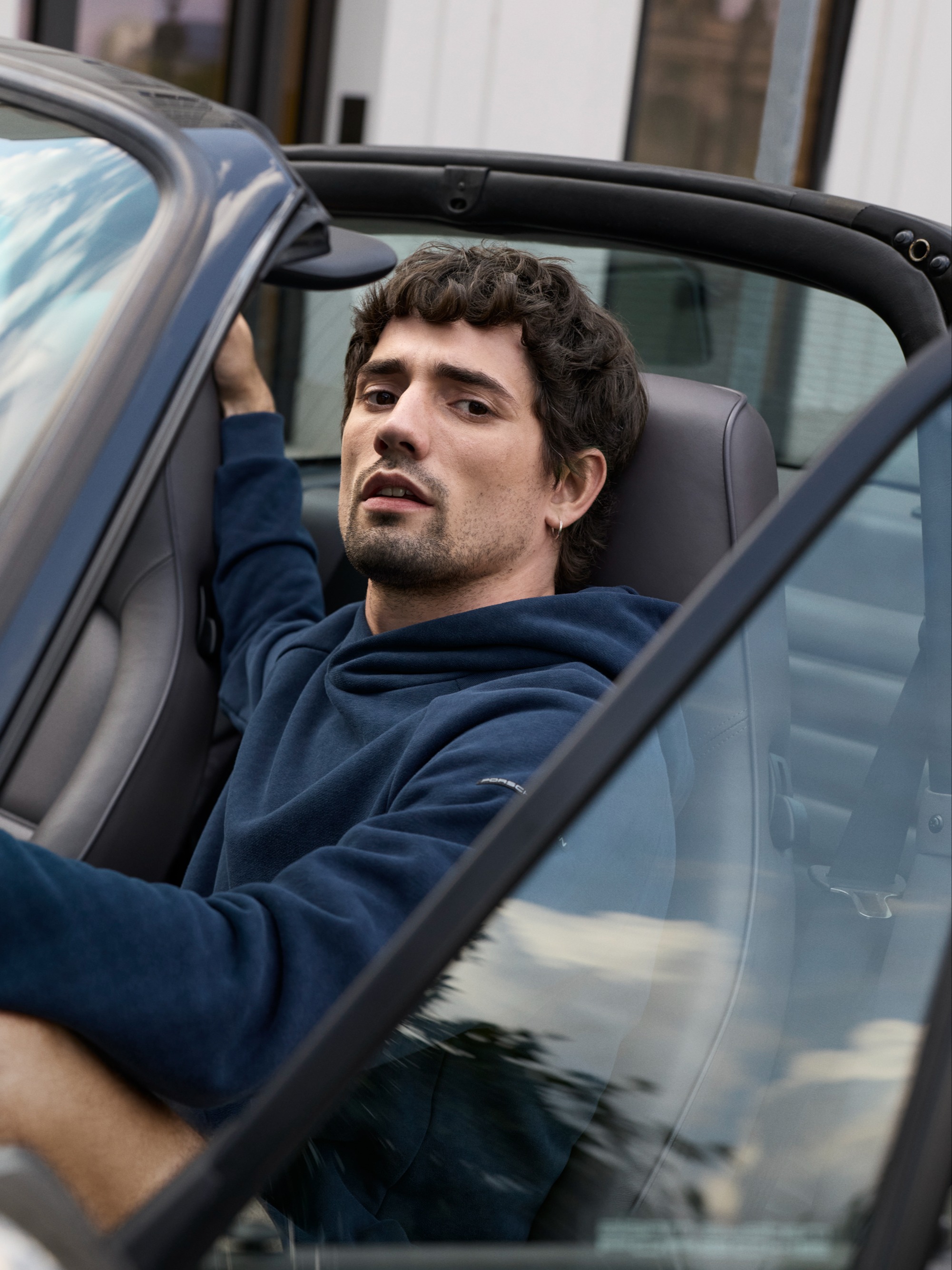 Homem com t-shirt Porsche junto a um carro clássico azul num espaço interior iluminado pelo sol, com a mão apoiada no tejadilho, a usar boné e jeans.