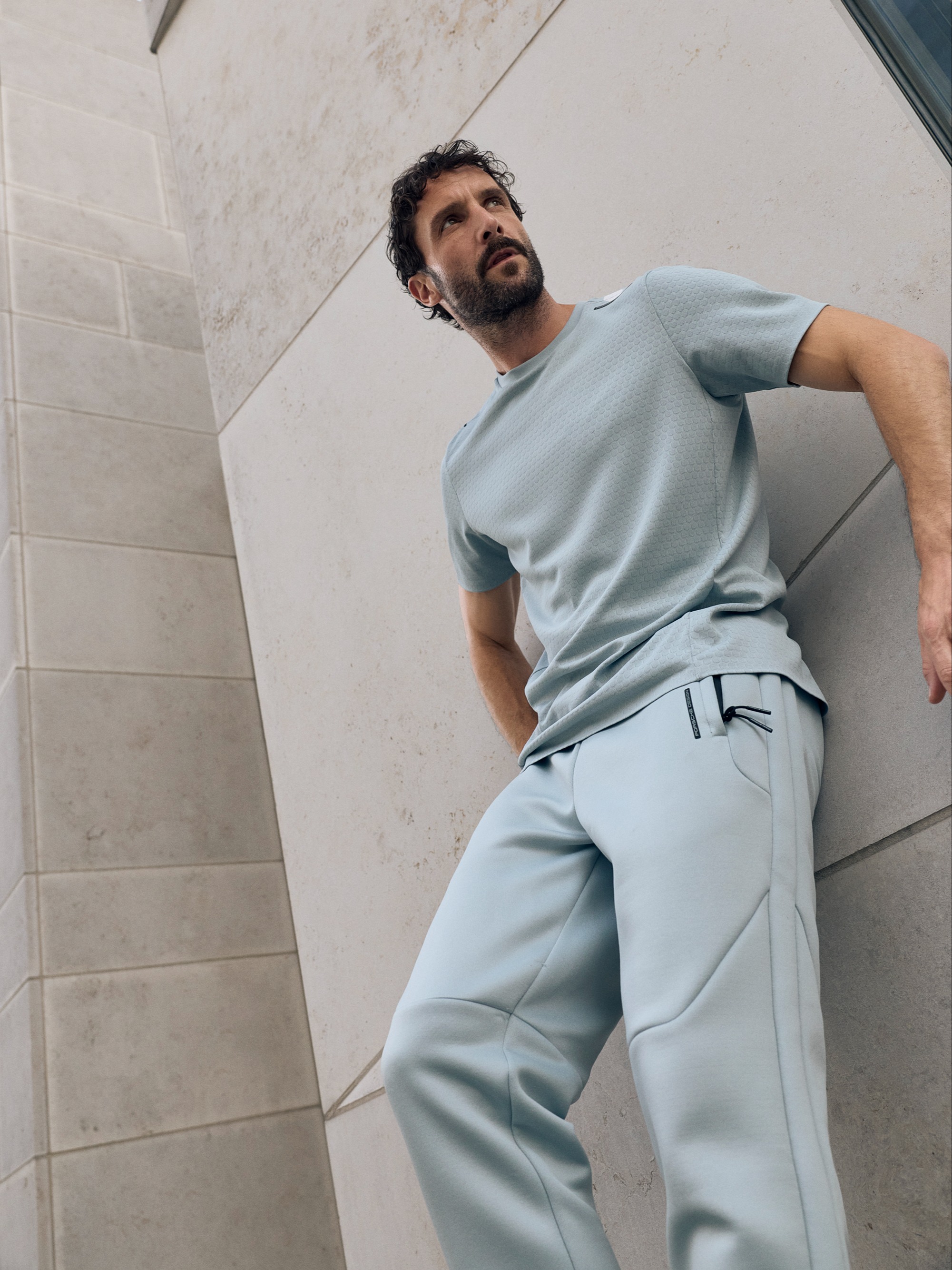 Man in light blue athletic wear leaning against a stone wall, looking upward with a thoughtful expression.