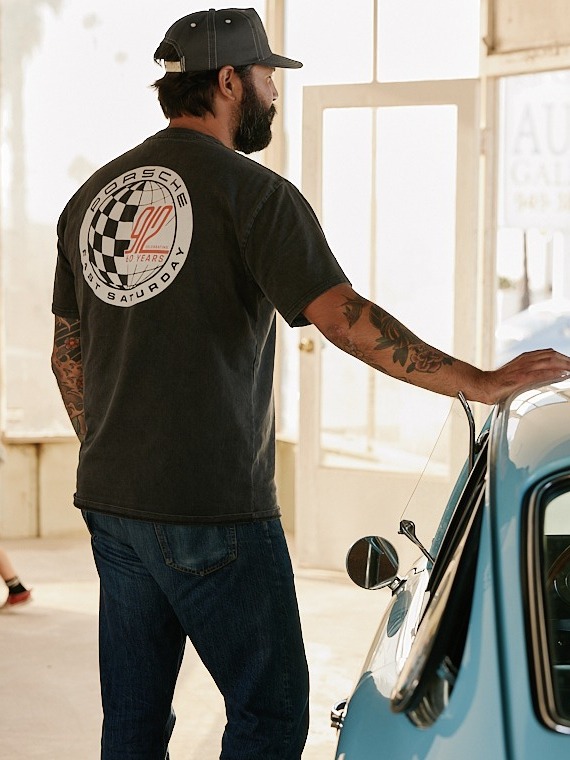 Man in Porsche shirt stands by a vintage blue car in a sunlit room, hand resting on the roof, wearing a cap and jeans.