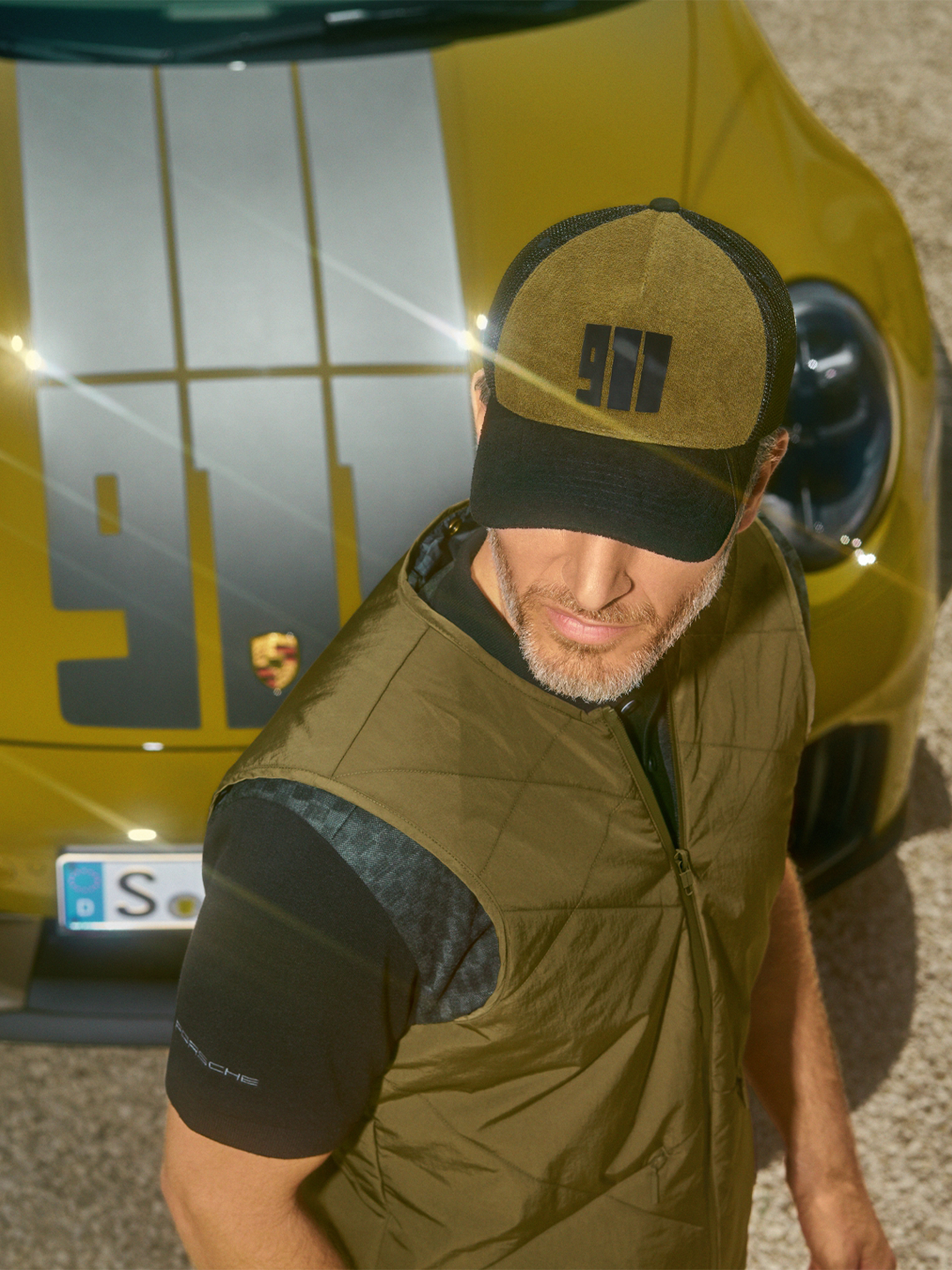 Man in Porsche shirt stands by a vintage blue car in a sunlit room, hand resting on the roof, wearing a cap and jeans.