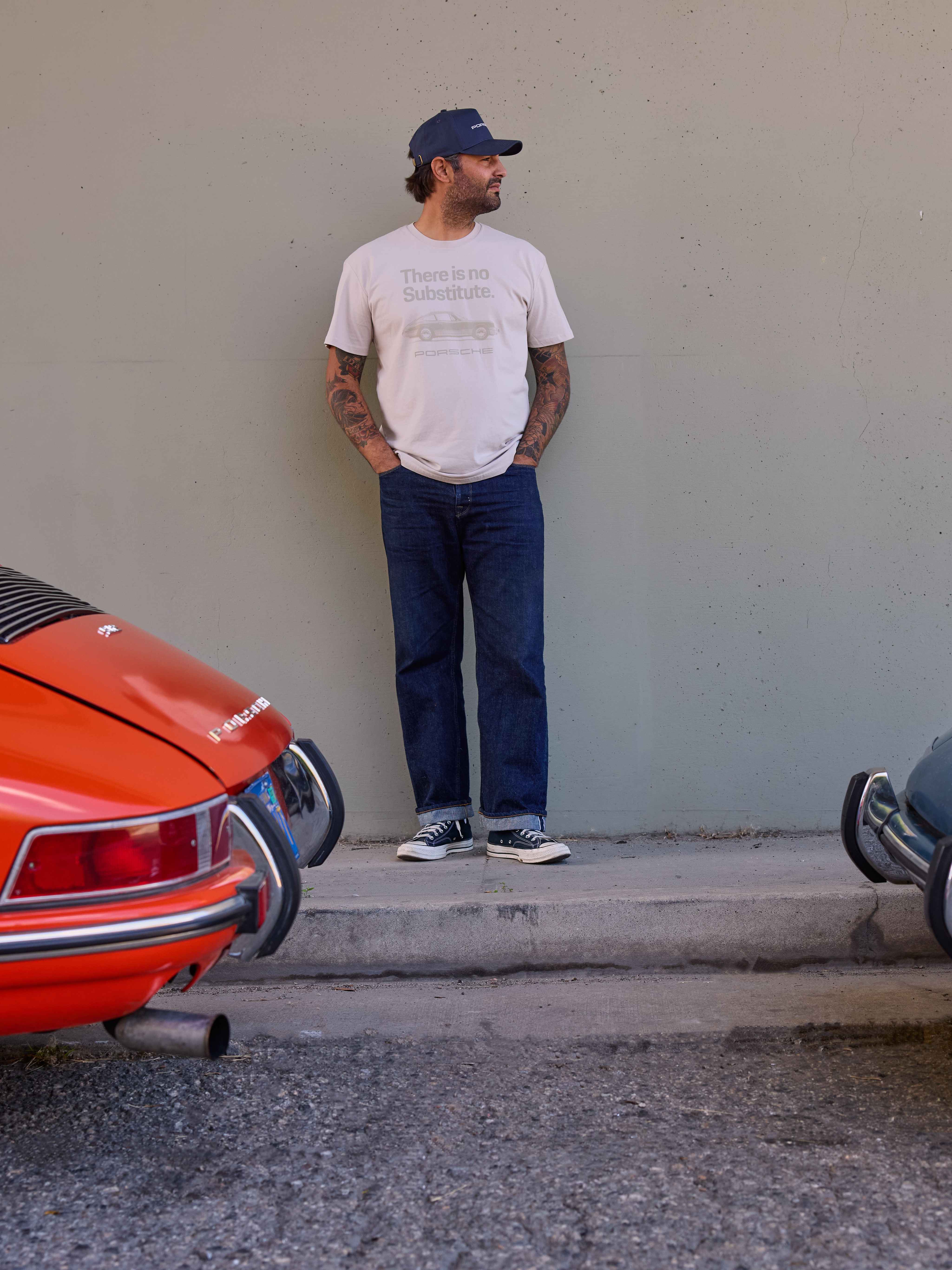 A man wearing a navy cap, a beige Porsche T-shirt and jeans is leaning against a grey wall, between two Porsche rear ends.
