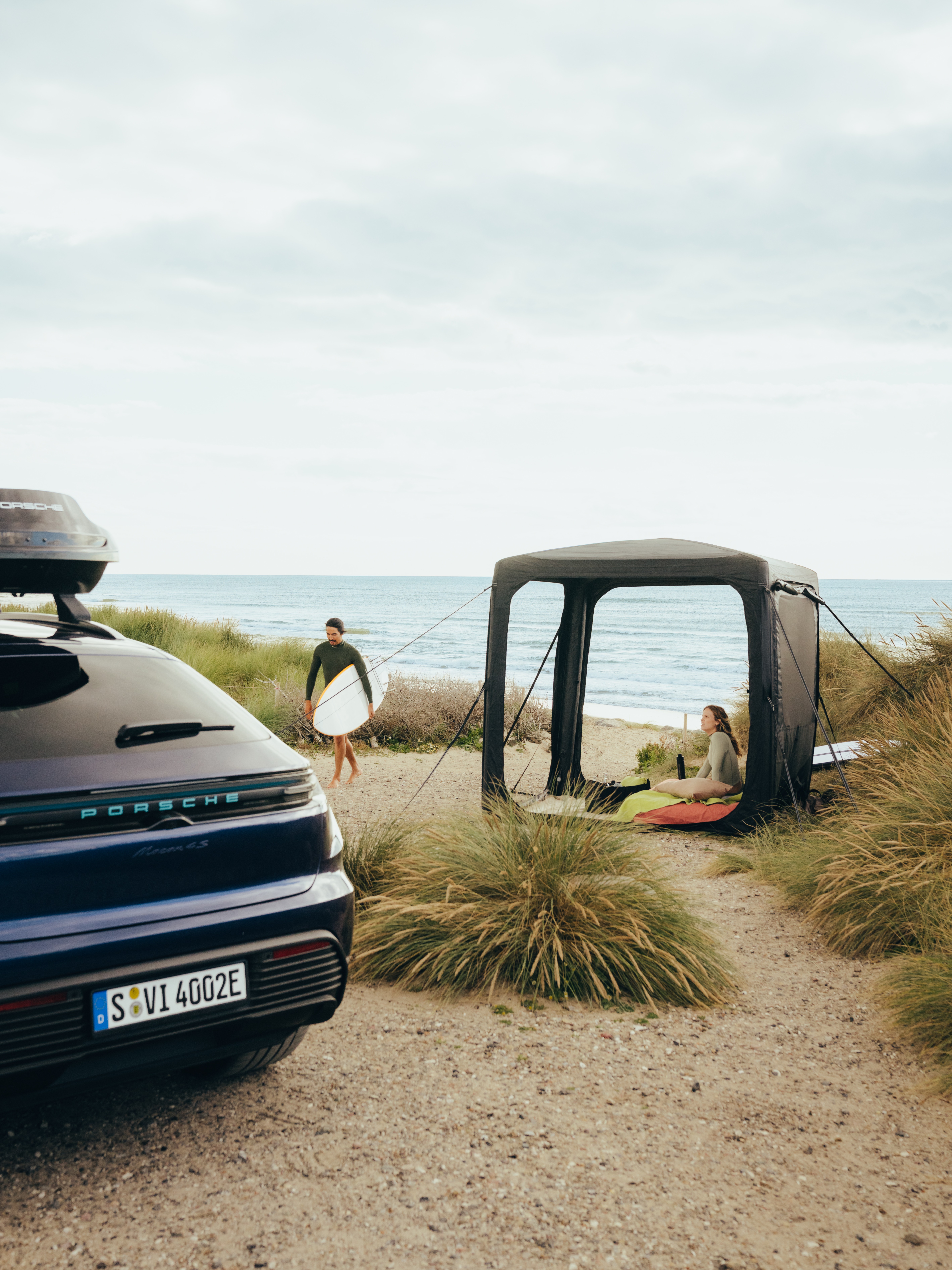 Porsche-SUV mit Dachbox, geparkt neben Dünen; Surfer mit Surfbrett geht an einem schwarzen Strandzelt am Meer vorbei.