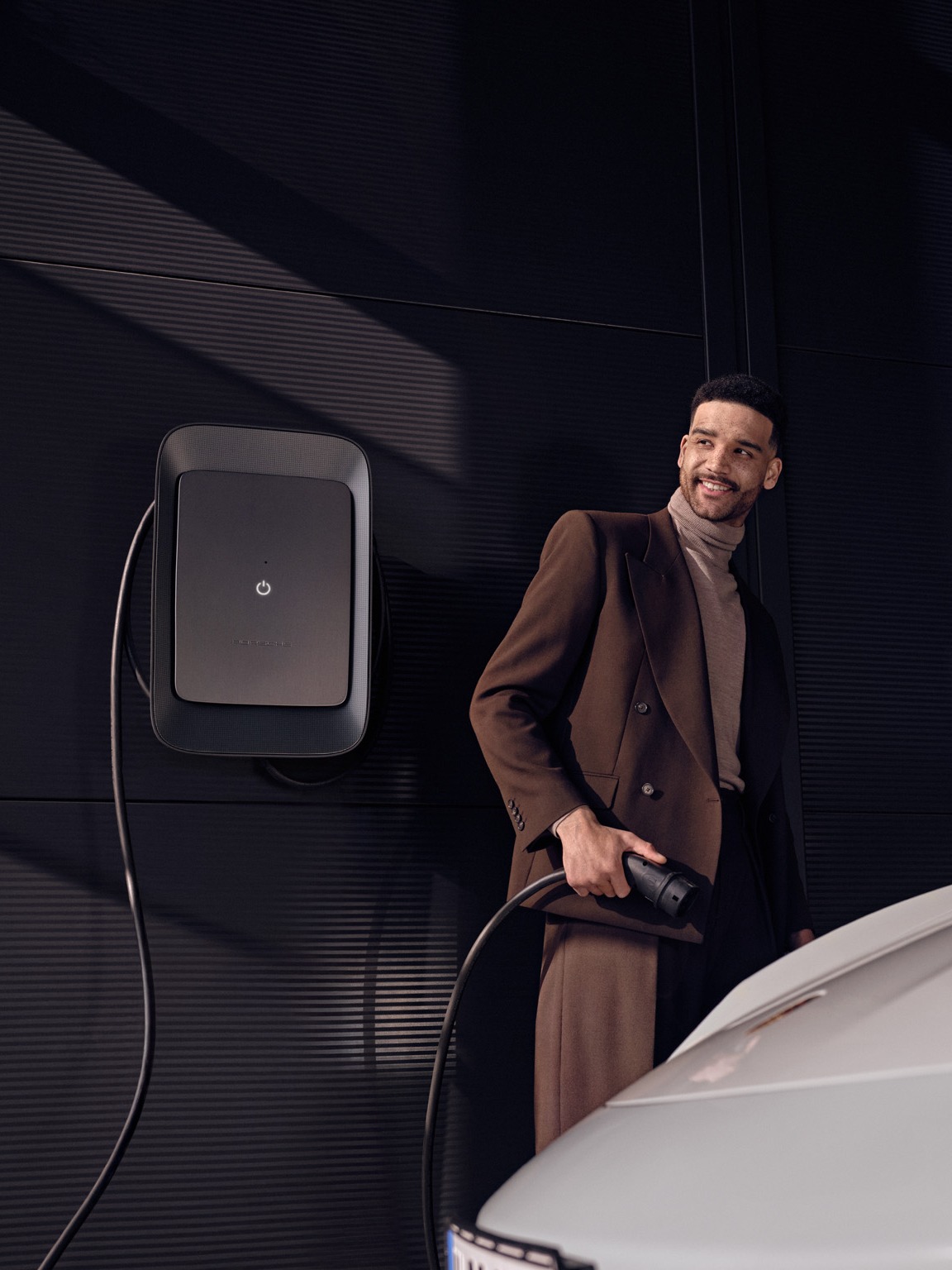 Man in brown suit charging a white car with a wall-mounted charger, dark background.