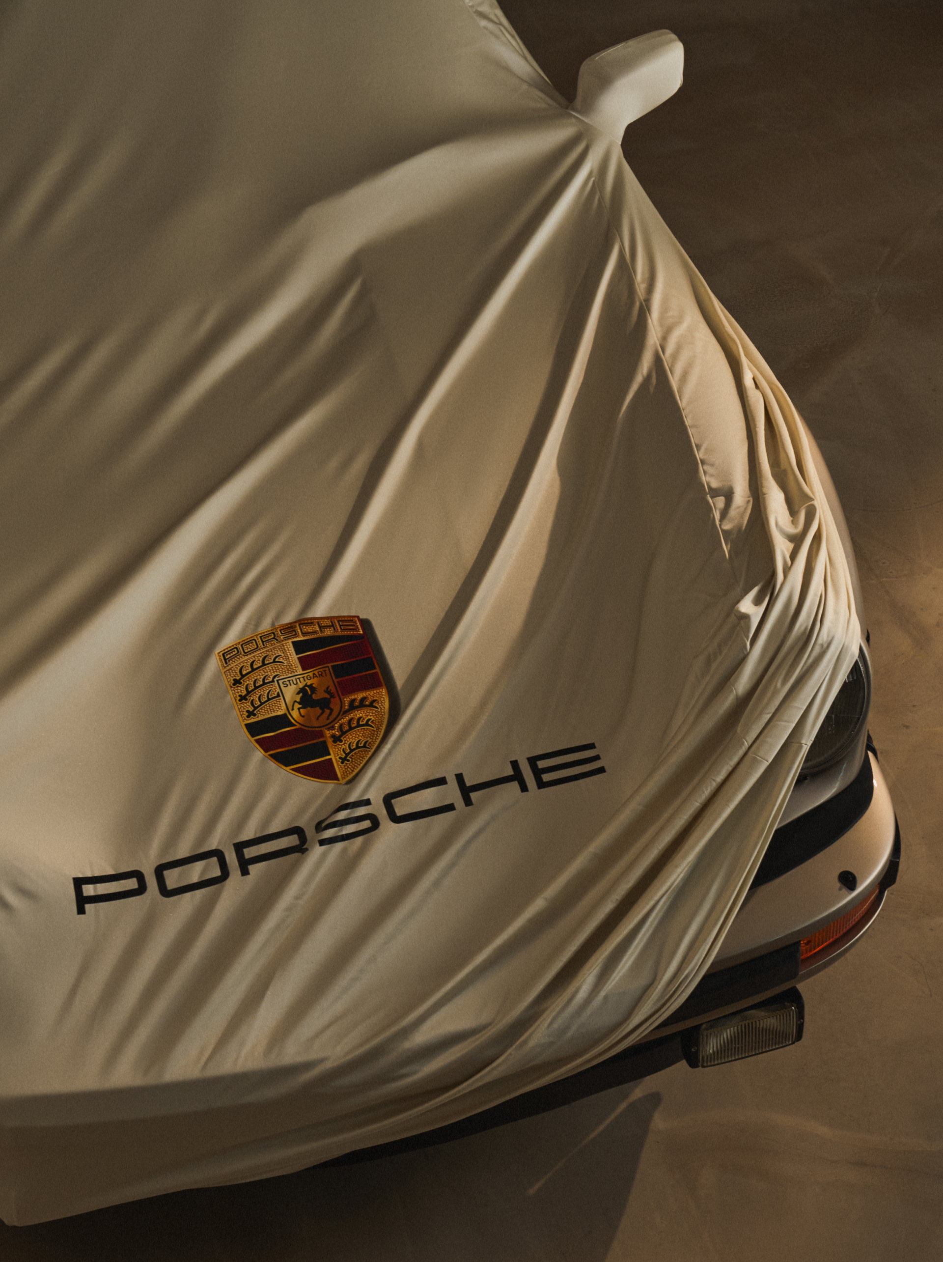 Car covered with beige fabric, displaying Porsche logo and text, partially revealing front headlight and bumper.