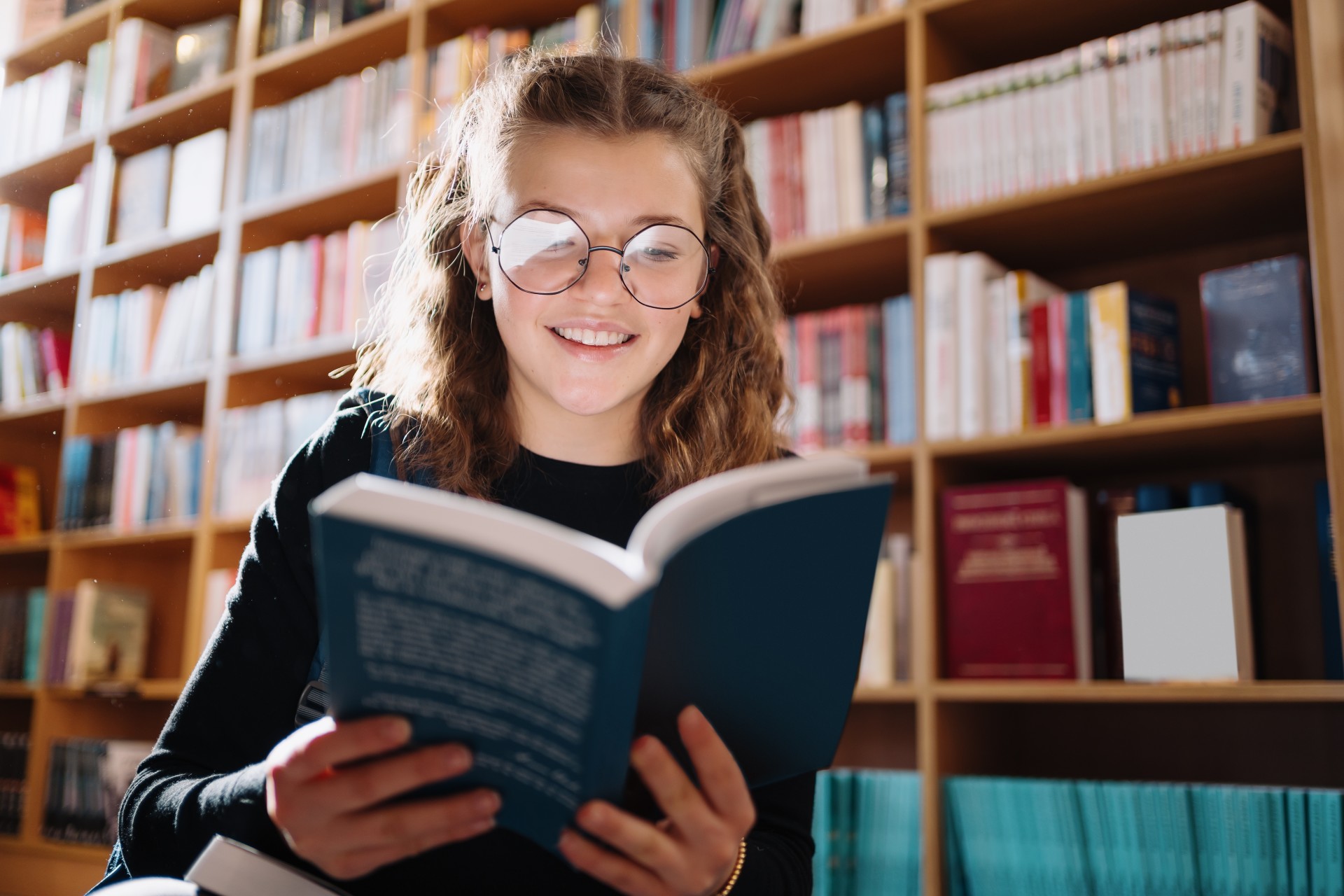 Młoda dziewczyna stoi przed ragałami pełnymi książek. Jedną książkę trzyma otwartą i czyta ją z uśmiechem na twarzy.