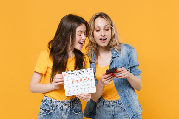 Dwie młode dziewczyny rozmawiają ze sobą i patrzą razem w telefon komórkowy. Jedna z nich trzyma również kalendarz.