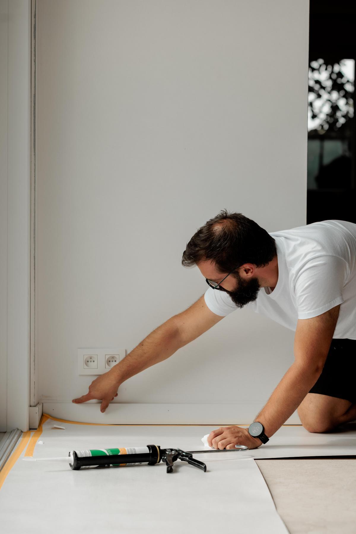 Een man in een wit shirt brengt kit aan op de onderkant van een witte muur in de buurt van een stopcontact, met behulp van een kitpistool en wijzend langs de rand van de muur.