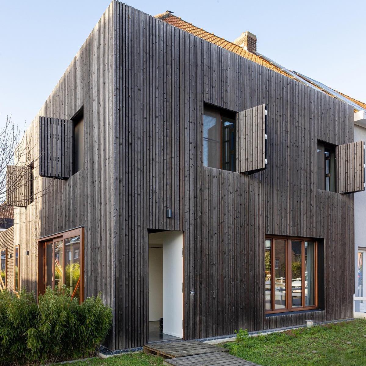 Une maison moderne à deux étages avec une façade verticale en bois foncé, de grandes fenêtres et des volets en bois ouverts à l'étage supérieur.