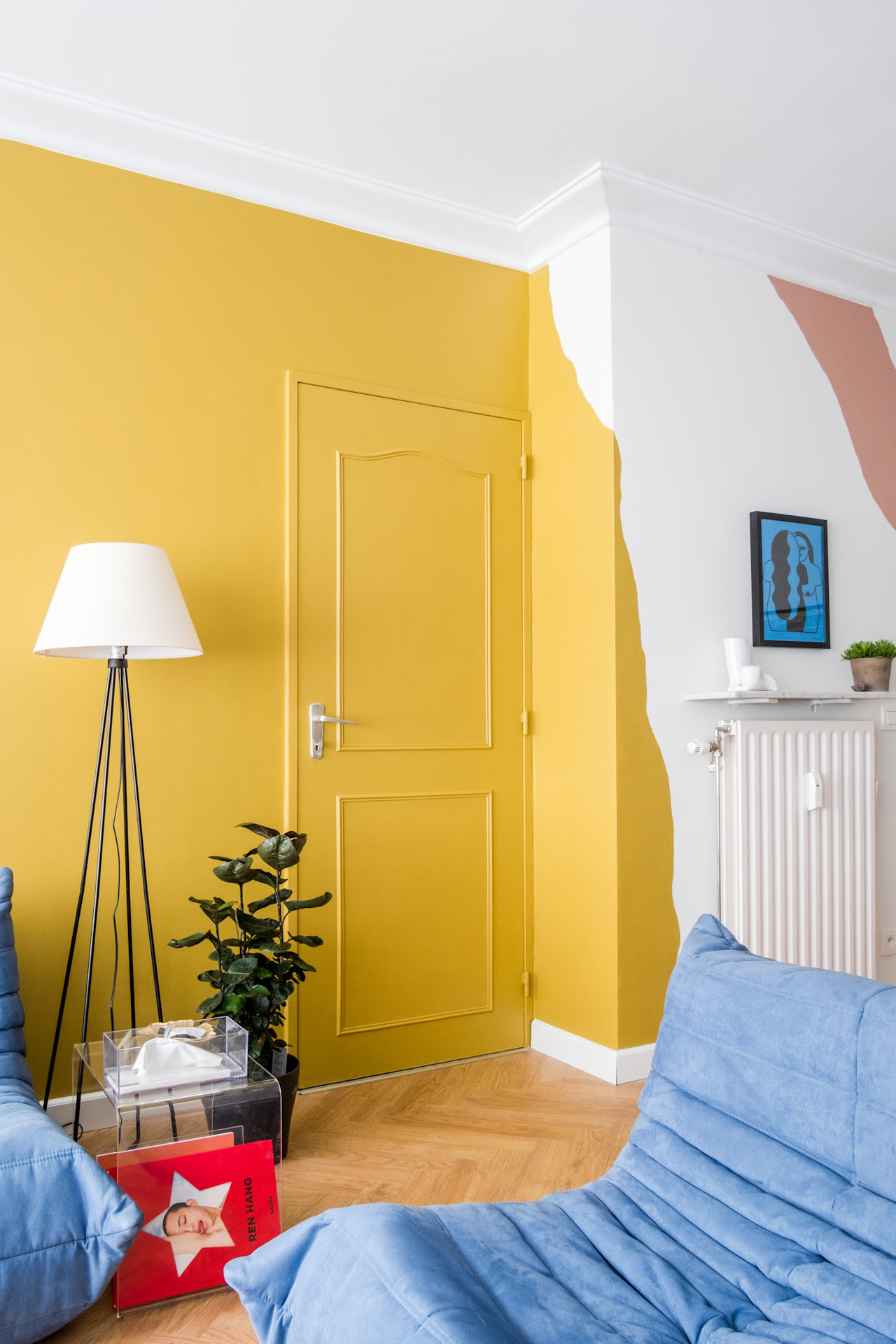 Un salon moderne avec un mur et une porte jaunes, des fauteuils bleus avec des coussins, un lampadaire blanc, une plante en pot, une petite table d'appoint transparente sur laquelle repose un livre rouge.