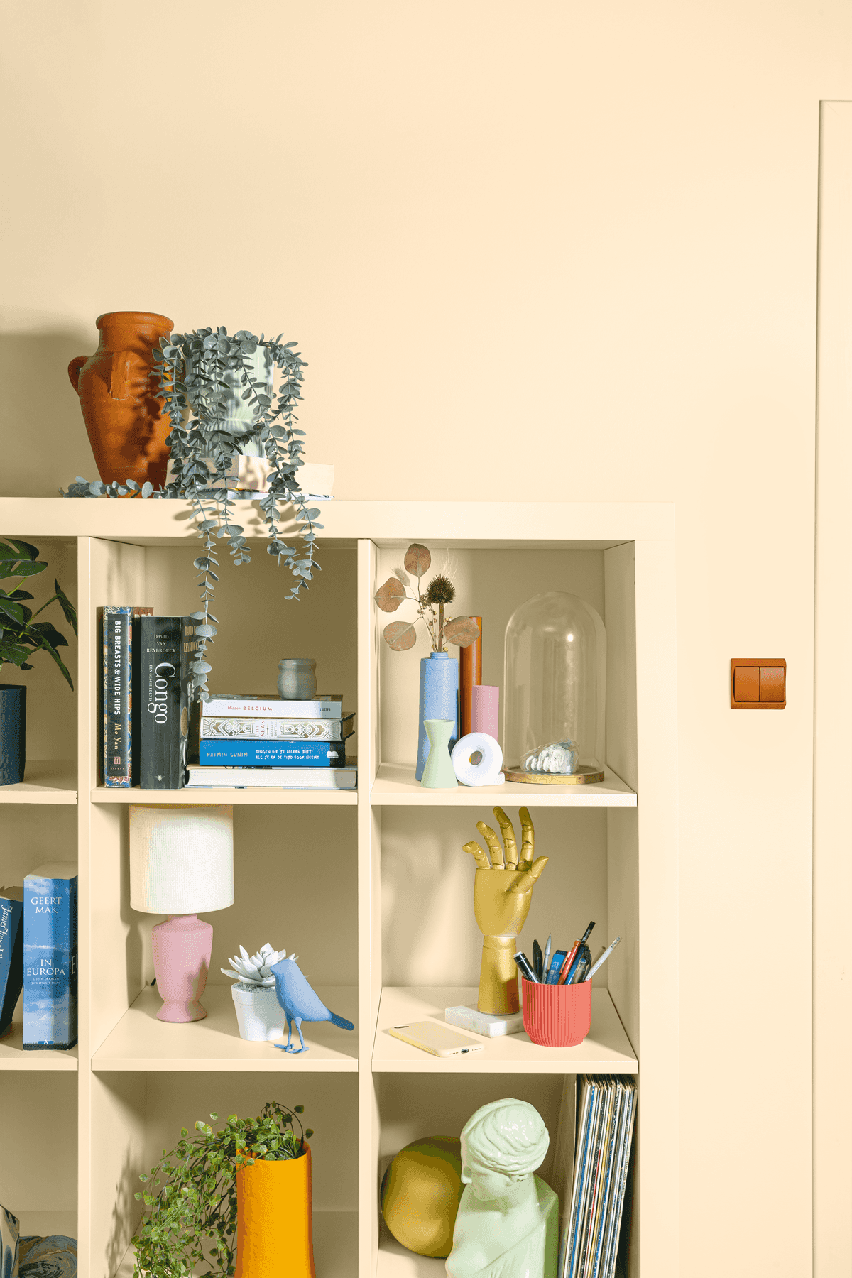 Op een beige schap staan boeken, planten, een keramische kan, een gouden handsculptuur, gekleurde bekers met pennen en decoratieve voorwerpen tegen een crèmekleurige muur met een bruine lichtschakelaar.