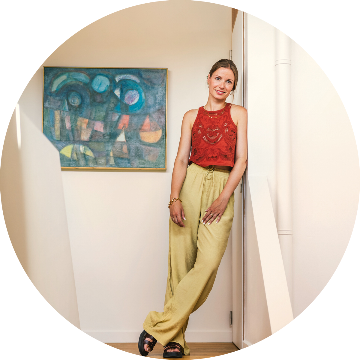 Een vrouw in een rode mouwloze top en gele broek staat glimlachend bij een witte muur, met een kleurrijk abstract schilderij op de achtergrond.