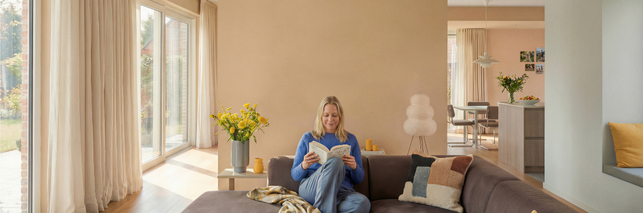 Een vrouw zit comfortabel op een bruine bank in een lichte, moderne woonkamer een boek te lezen. Grote ramen met lichte gordijnen laten natuurlijk licht binnen en gele bloemen sieren de salontafel. De keuken is zichtbaar op de achtergrond.