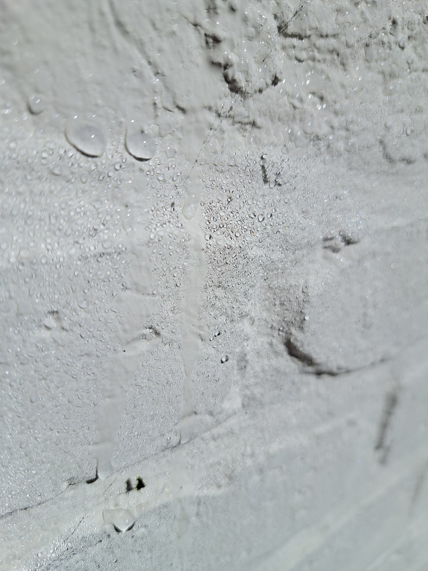Close-up van een witgeschilderde bakstenen muur met verschillende waterdruppels op het oppervlak, die de ruwe textuur van de muur en de ongelijkmatige verflaag benadrukken. De foto is onder een hoek genomen, waardoor een kleine scherptediepte ontstaat.