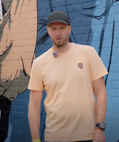 Een man in een lichtperzikkleurig T-shirt en een zwarte pet staat voor een kleurrijke muurschildering op een bakstenen muur. Hij draagt een horloge en een microfoon, en houdt één hand ontspannen langs zijn lichaam.