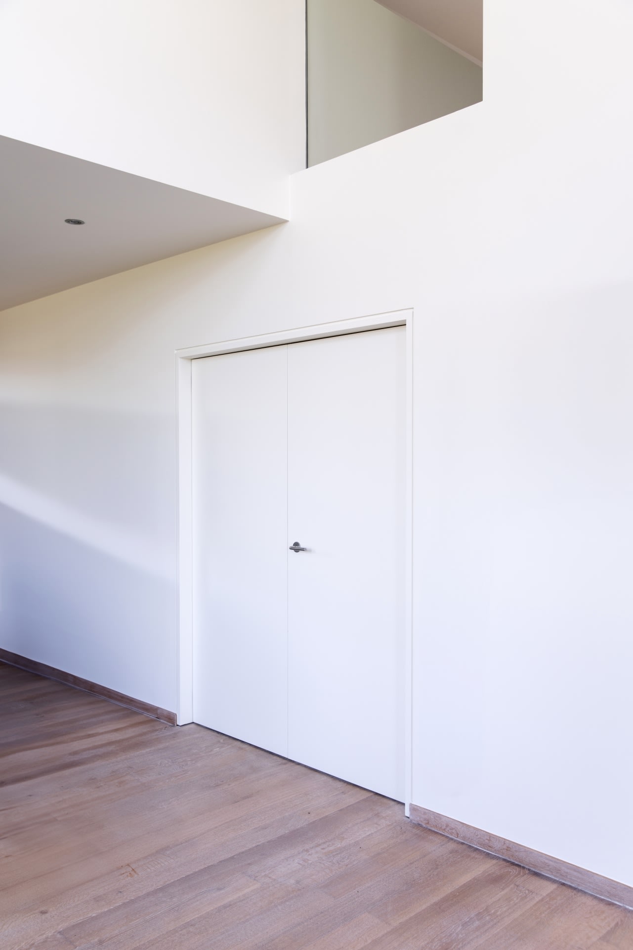Une double porte blanche avec des poignées argentées dans un mur blanc uni, à côté d'un sol en bois dans un intérieur moderne et minimaliste.