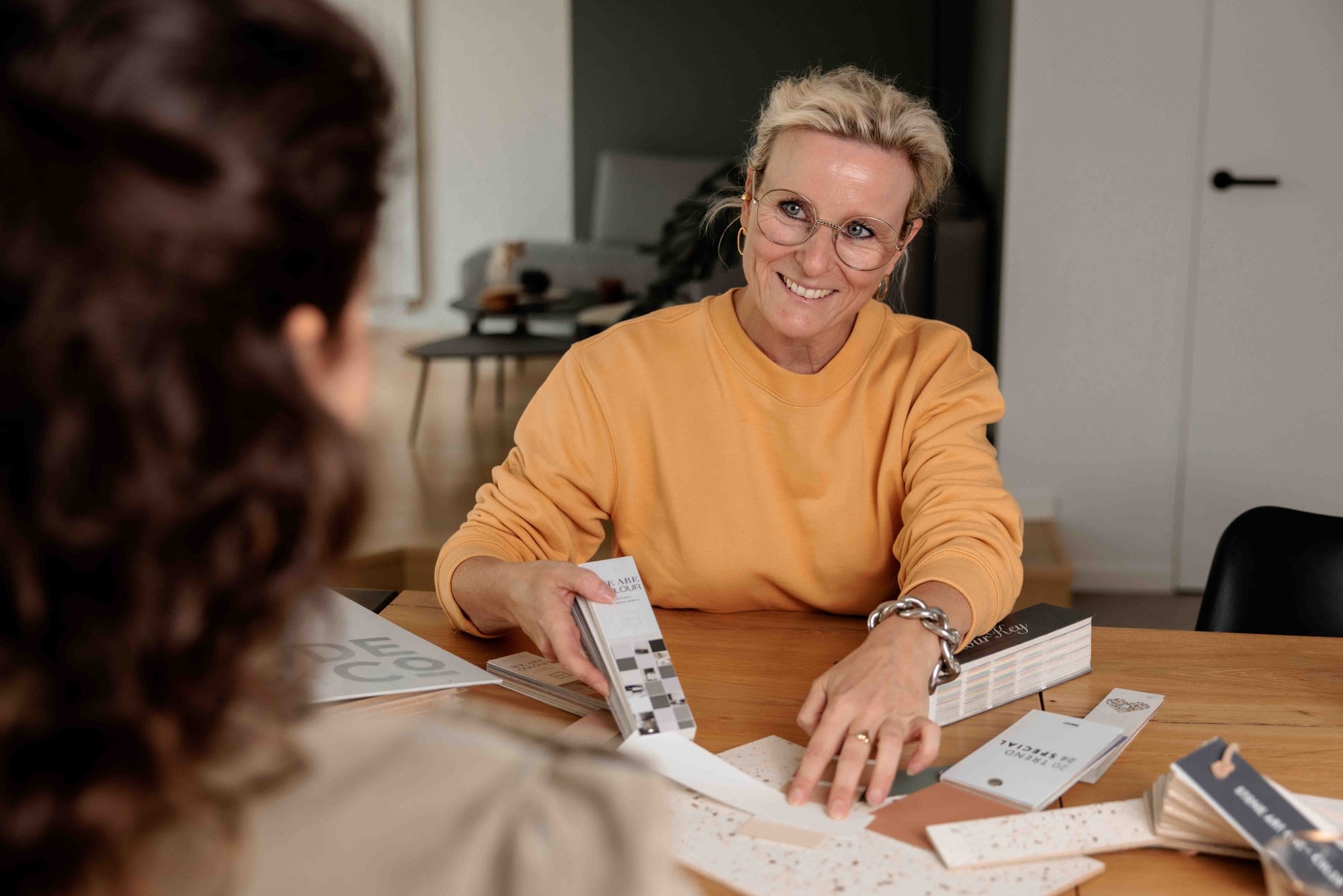 Een vrouw met bril en gele trui toont kleur- en materiaalstalen aan een andere persoon aan een tafel in een modern kantoor.